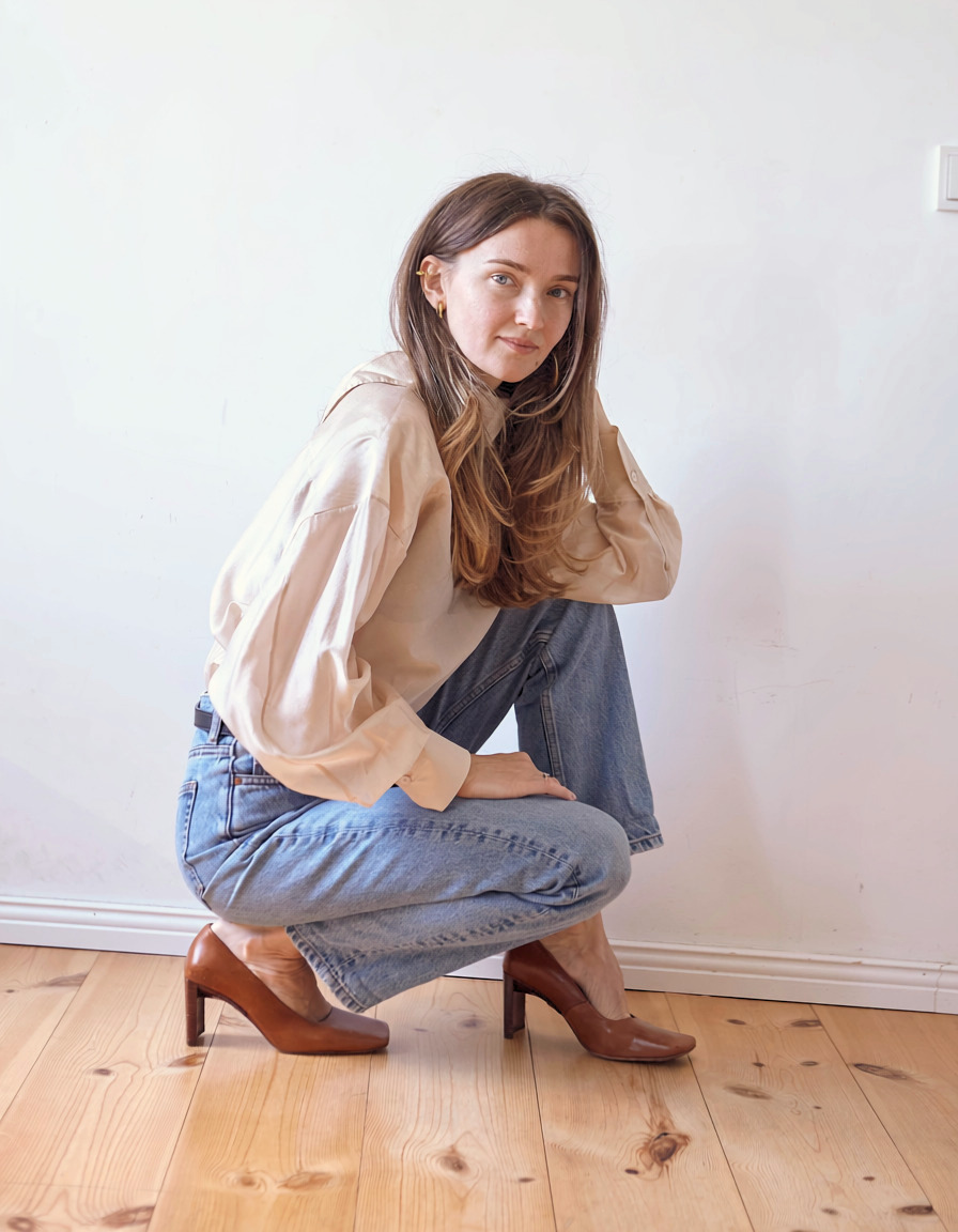 Woman crouching against a white wall in a beige blouse and denim